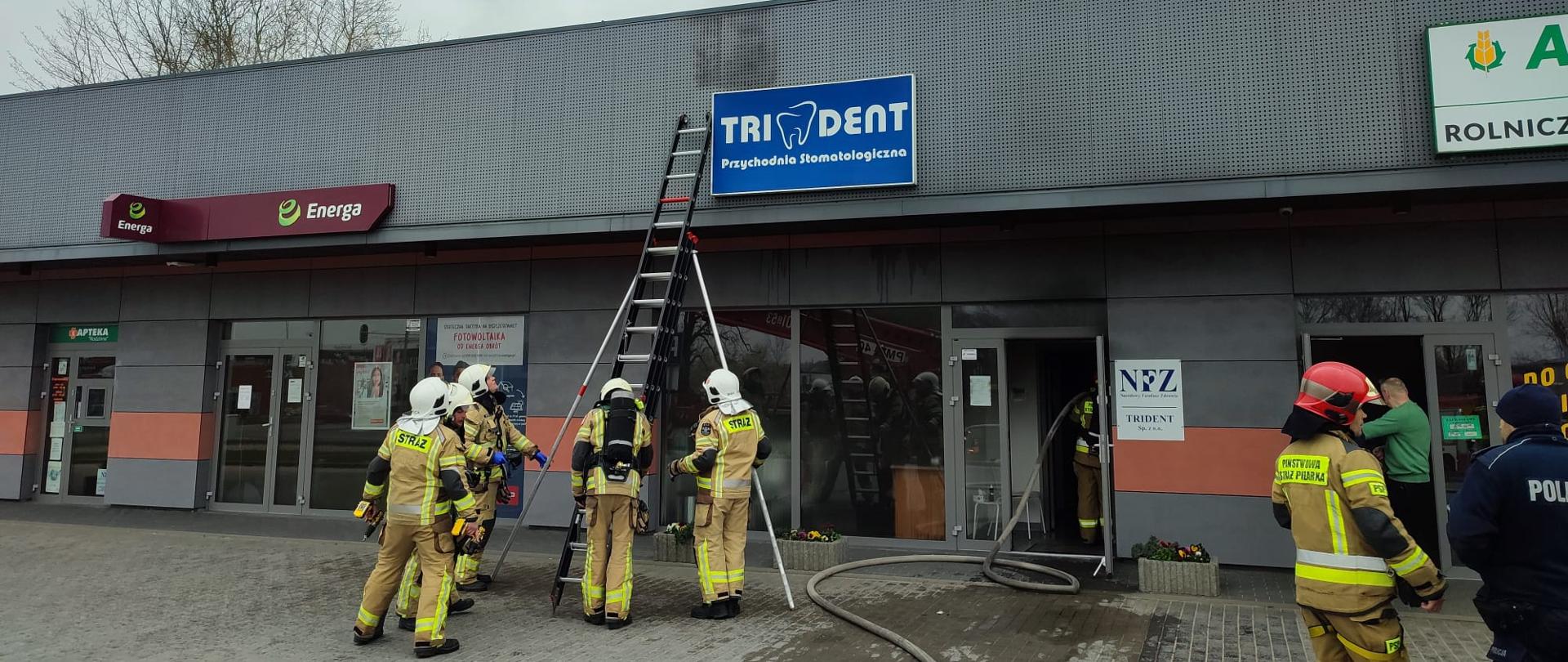 Zdjęcie przedstawia strażaków ubranych w ubrania specjalne koloru żółtego. Ratownicy stoją przy rozstawionej drabinie przystawionej do budynku. W tle widać pomieszczenie objęte działaniami ratowniczymi.