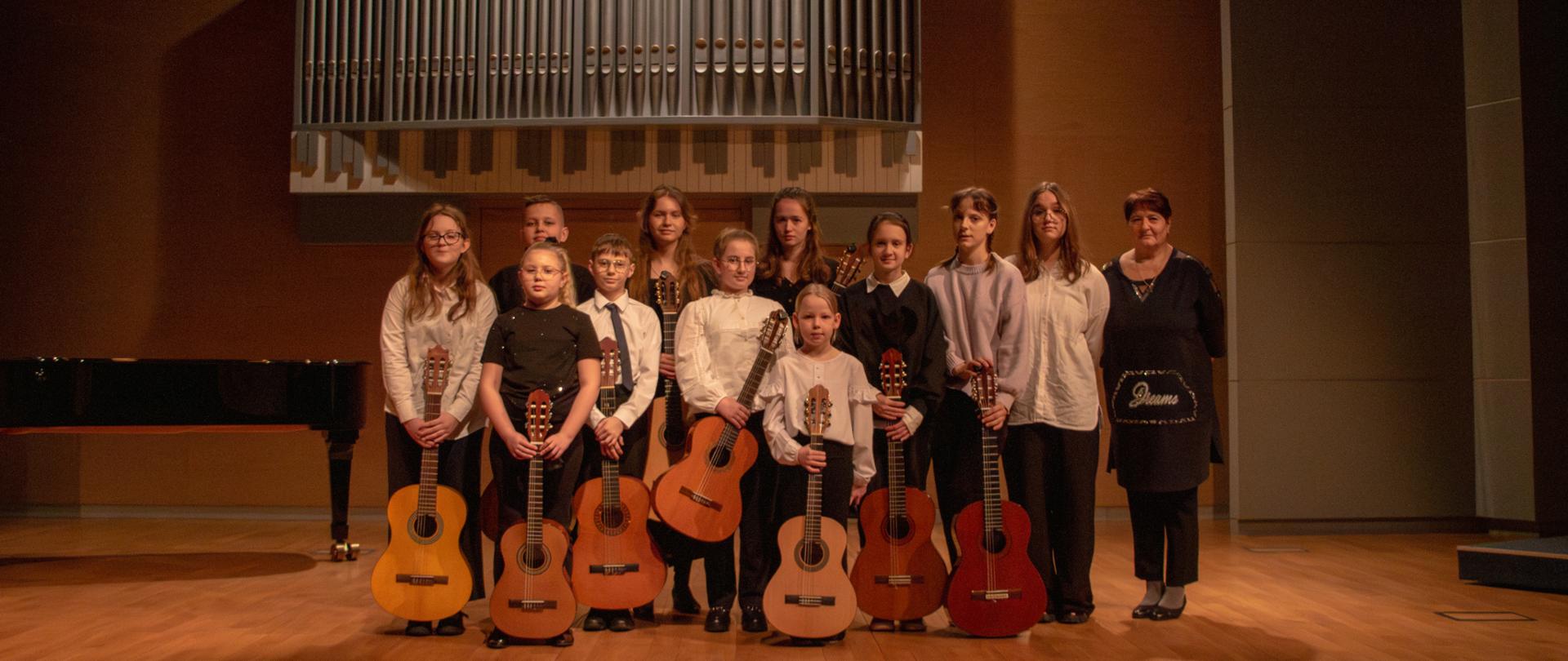 Zdjęcie grupowe wykonane na scenie sali koncertowej po występie. W centrum znajduje się grupa dzieci i młodzieży (dziesięć osób) oraz jedna dorosła kobieta po prawej stronie. Większość uczniów trzyma przed sobą gitary klasyczne o różnych odcieniach drewna. Są ubrani galowo – dominują białe koszule, ciemne spodnie oraz ciemne swetry.
Tło i otoczenie:
Bezpośrednio za grupą, na ścianie, zawieszone są duże organy piszczałkowe o srebrzystych rurach ułożonych asymetrycznie.
Po lewej stronie fragmentu kadru widoczny jest czarny, błyszczący fortepian skrzydłowy.
Scena ma jasną, drewnianą podłogę, a ściany są w odcieniach ciepłego brązu i szarości. Oświetlenie jest nastrojowe i równomierne.