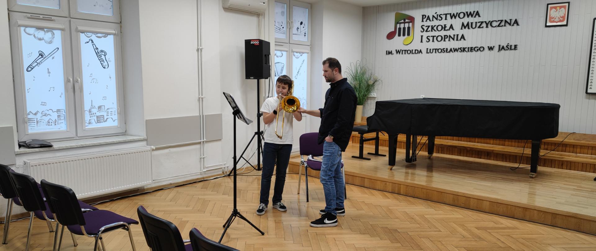 Mężczyzna w czarnej koszuli prawą ręką trzyma lewą rękę chłopca grającego na puzonie obaj patrzą na stojący przed nimi pulpit z nutami. Za mężczyzną w czarnej koszuli stoi krzesło na którym leży srebrna trąbka. Za krzesłem stoi czarny fortepian. W tle ściana w jasnym kolorze z napisem państwowa szkoła muzyczna pierwszego stopnia im Witolda Lutosławskiego w Jaśle obok napisu logo szkoły przedstawiające dwie kolorowe nutki, między którymi widać trzy klawisze fortepianu.