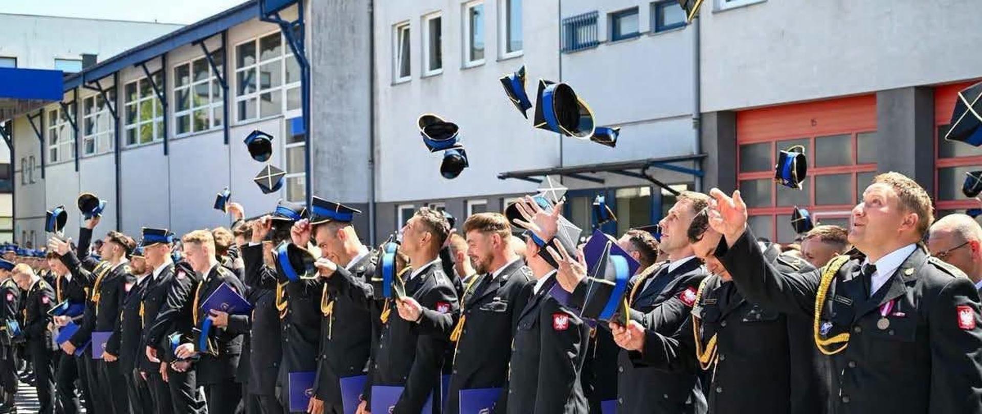 Duża grupa strażaków w mundurach galowych ustawionych w szeregu podrzuca czapki na koniec akademii wręczenia dyplomów ukończenia szkoły i nadania stopni. W tle budynek szkoły.