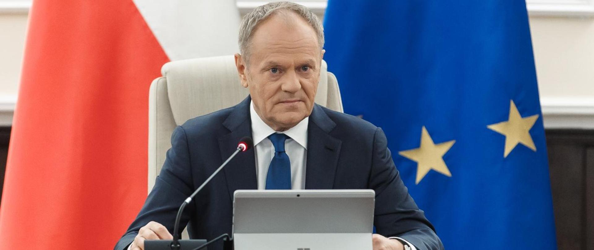 Premier Donald Tusk przemawiający przed posiedzeniem Rady Ministrów, na tle flag Polski i Unii Europejskiej
