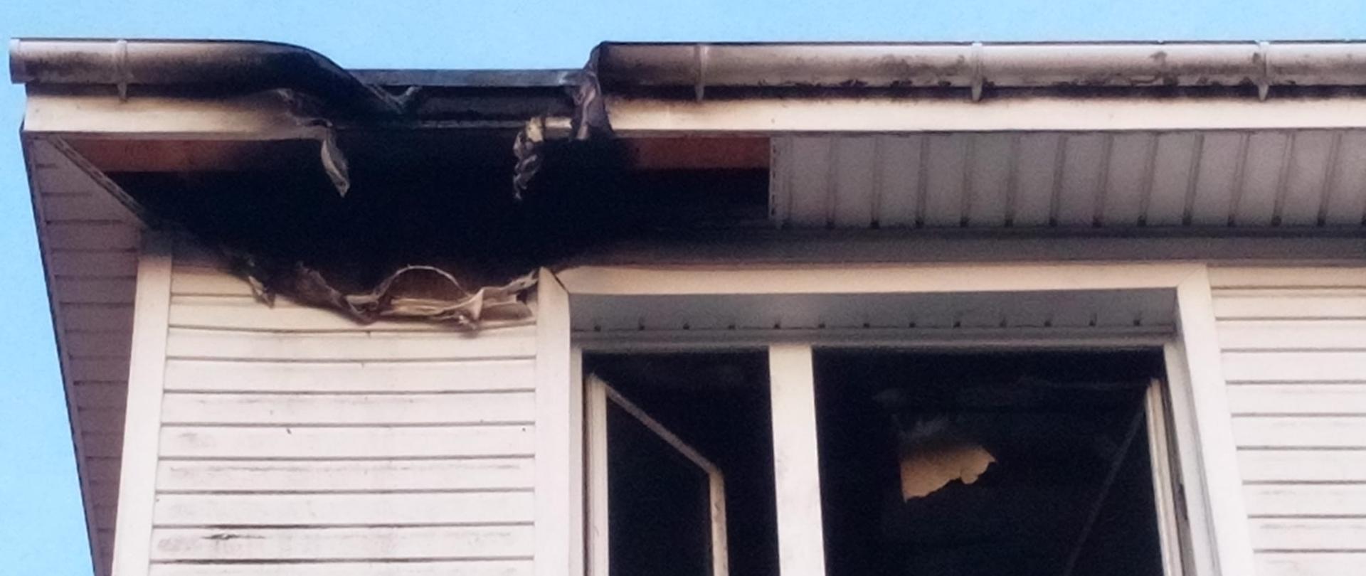 Na zdjęciu widoczne okno na poddaszu z widoczną nadpaloną dziurą wychodzącą na dach. Elewacja w kolorze białym.