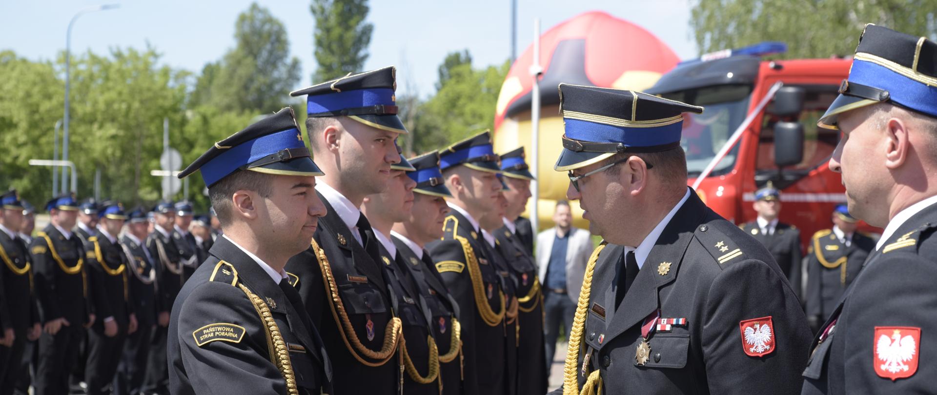 Na zdjęciu widzimy strażaków stojących w szeregu. Strażacy ubrani są w galowe mundury koloru czarnego. Przed strażakami stoi przodem kolejnych dwóch strażaków. Strażak po prawej stronie podaje rękę jednemu strażakowi stojącemu w szeregu. W tle widzimy budynki mieszkalne, zielone drzewa a także wóz strażacki koloru czerwonego.