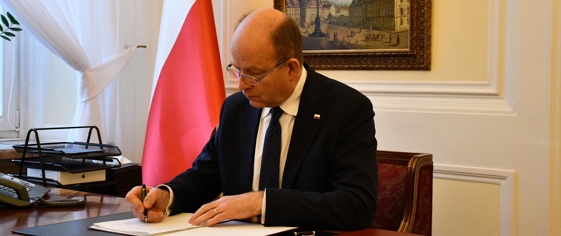 Na pierwszym planie zdjęcia znajduje się ubrany w granatowy garnitur, białą koszulę i ciemny krawat mężczyzna. Siedzi on za biurkiem, podpisując porozumienie w sprawie współdziałania w zakresie organizacji ratownictwa medycznego. W tle znajduje się biało-czerwona flaga oraz wiszący przy ścianie obraz przedstawiający warszawski plac Zamkowy z Zamkiem Królewskim i Kolumną Zygmunta. 