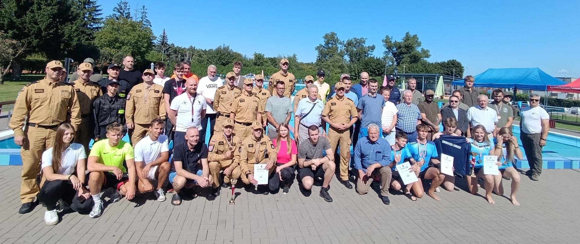 Wspólne pamiątkowe zdjęcie, kończące zawody pływackie na tle basenu. Są obecni przedstawiciele wszystkich służb mundurowych, w tym Leśników, policjantów, strażaków Państwowej Straży Pożarnej i Ochotniczej Straży Pożarnej, młodzieży szkolnej, Wodnego Ochotniczego Pogotowia Ratowniczego oraz Władz samorządowych i zaproszonych gości.
