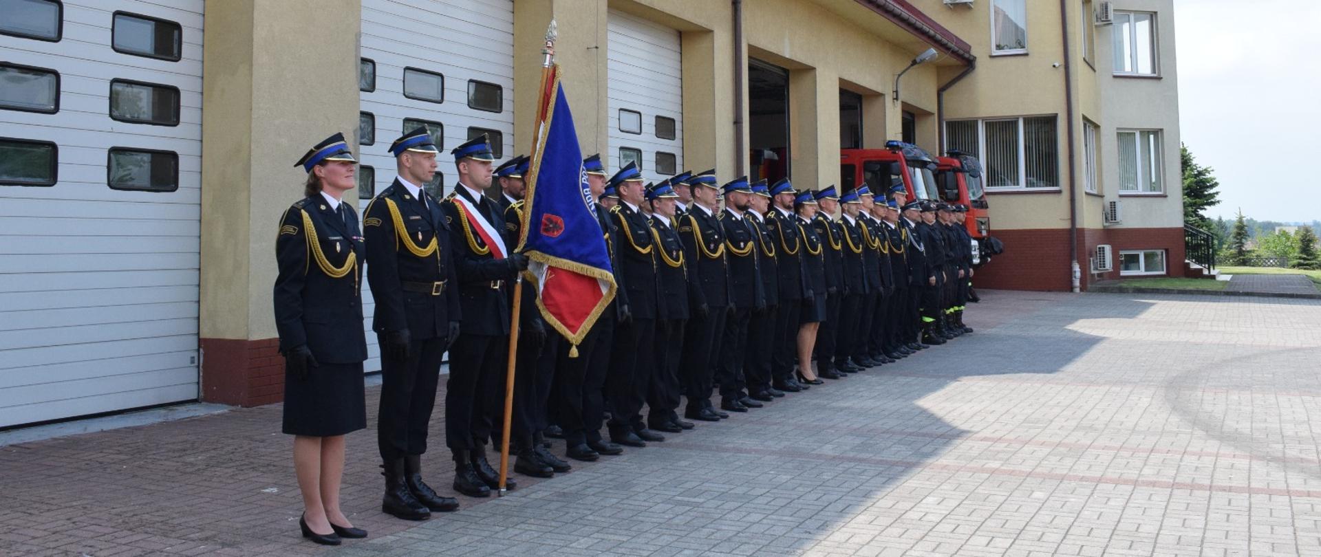 Pododdział stoi w strojach galowych w dwurzędzie. Na początku jeden z strażaków trzyma sztandar komendy skierowany do przodu. 