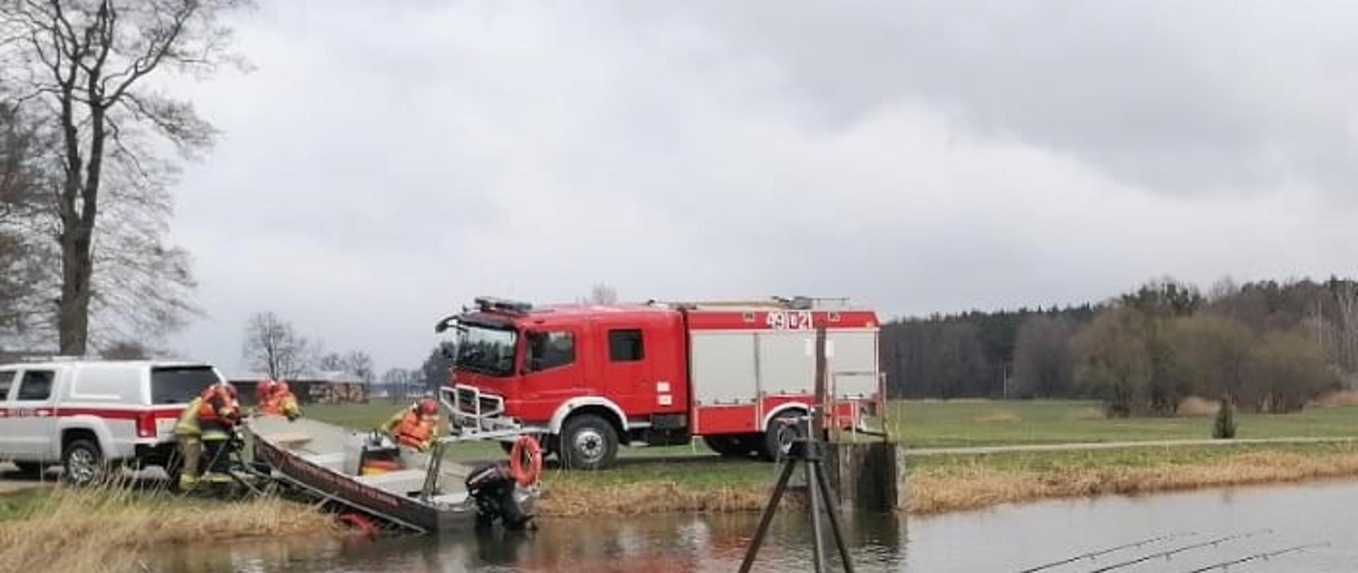 Zdjęcie przedstawia staw, dwa samochody pożarnicze i czterech strażaków podczas wodowania łodzi. na pierwszym planie jest pomost na którym stoją dwie wędki.