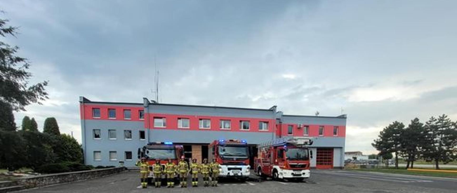 Na zdjęciu znajdują się samochody strażackie ustawione przed budynkiem komendy oraz strażacy oddający honor