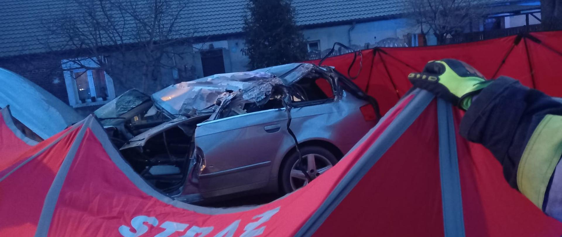 Widać rozstawiony czerwony parawan, za nim uszkodzony srebrny samochód od strony kierowcy