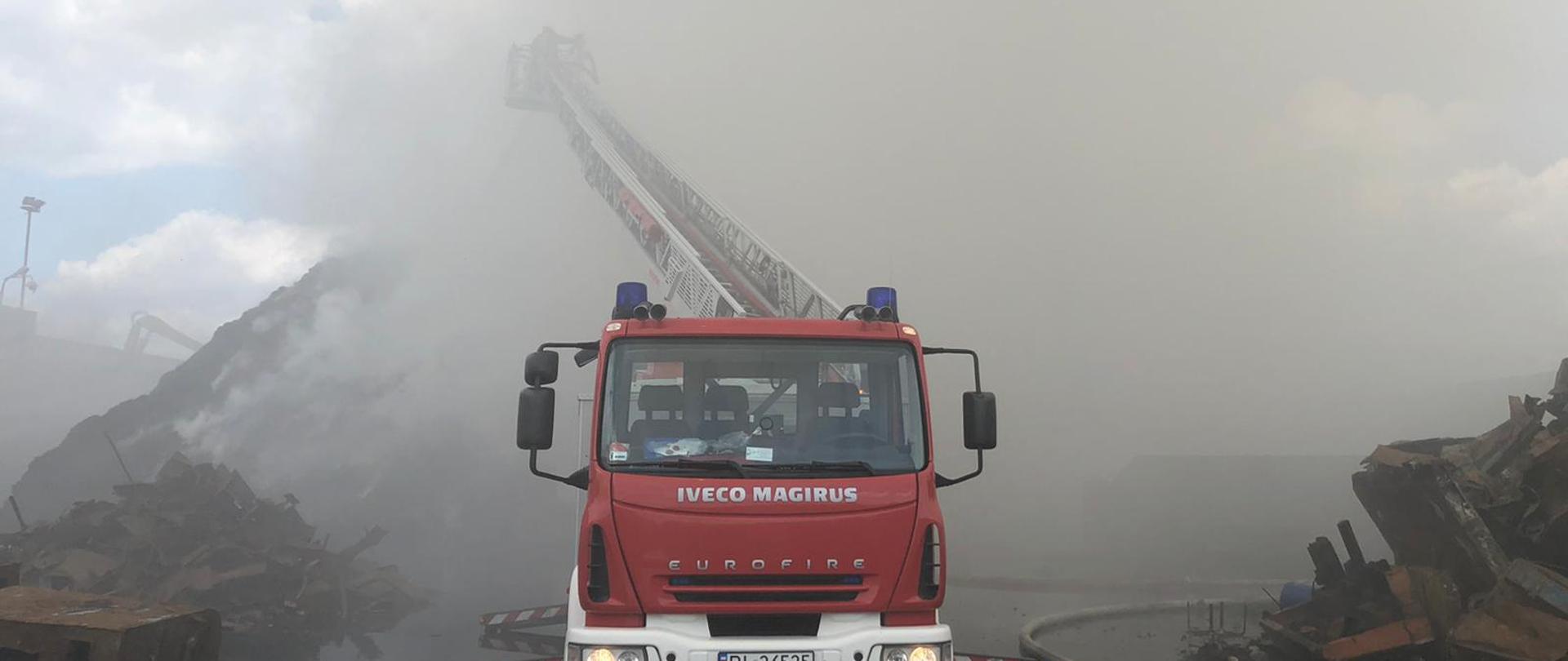 Strażacka drabina mechaniczna stoi na drodze z wysuniętą drabiną. Na ziemi leżą węże strażackie. W tle widoczna dymiąca hałda. 