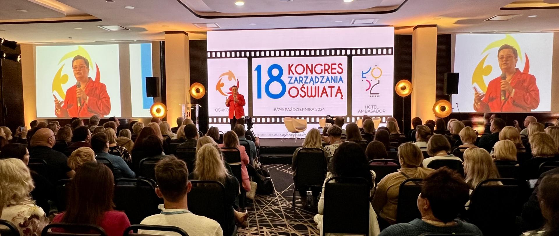 Wiceminister Lubnauer stoi na scenie i trzyma w ręku mikrofon. W tle znajduje się duży ekran, na którym wyświetlane są informacje o wydarzeniu. Na ekranie w widocznym miejscu widnieje napis "18 KONGRES ZARZĄDZANIA OŚWIATĄ" oraz daty "6/7-9 PAŹDZIERNIKA 2024". PO bokach dwa duże telebimy. Przed sceną na w rzędach ustawianych krzesłach siedzą ludzie.