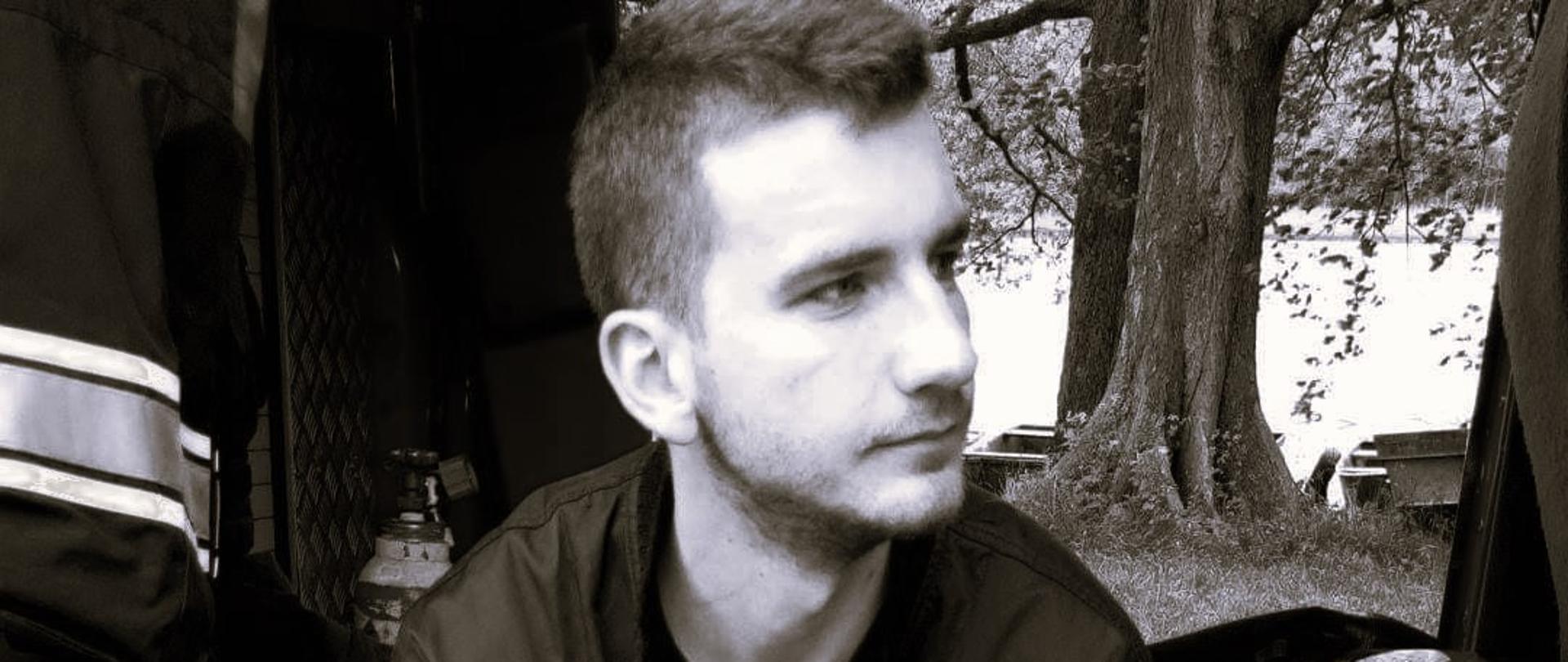 Czarno-białe zdjęcie przedstawiające strażaka w ubraniu strażackim siedzącego na progu samochodu.