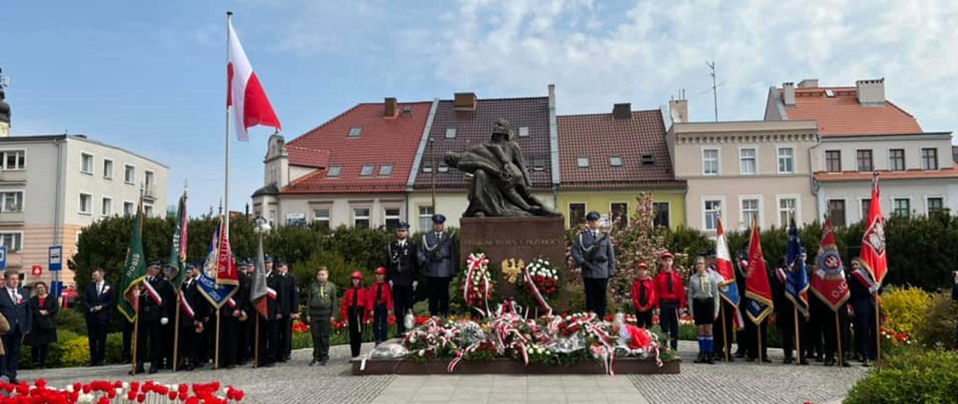 Na zdjęciu znajdują się poczty sztandarowe, warta honorowa przy pomniku, pomnik pod którym są złożone wiązanki kwiatów na tle budynków