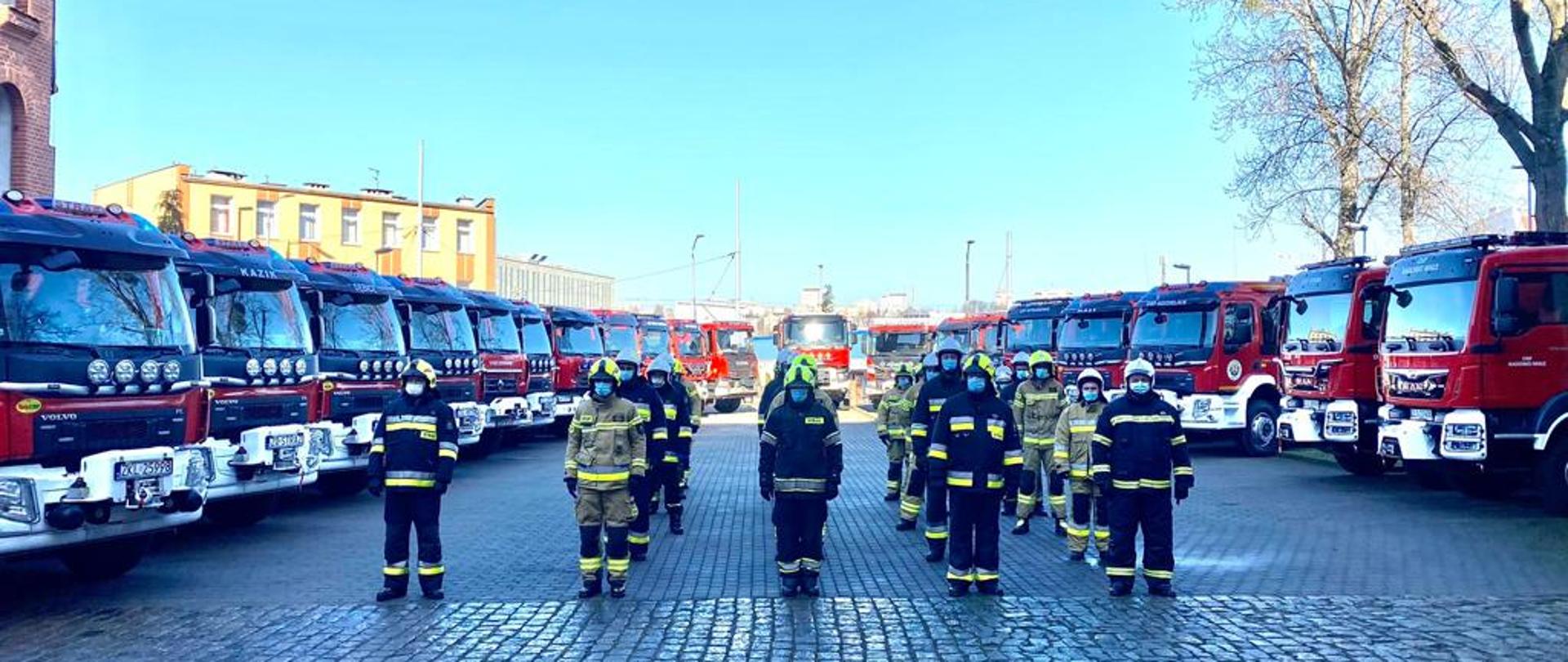 Na zdjęciu kilkunastu strażaków w umundurowaniu bojowym i w hełmach stoi w kilku rzędach między dwoma rzędami nowych samochodów ratowniczo-gaśniczych.