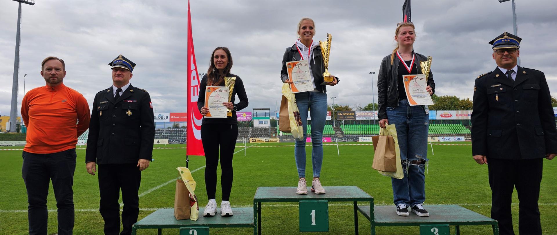 Zawodniczki stojące z pucharami dyplomami i medalami na podium. Obok nich strażacy w mundurach galowych.