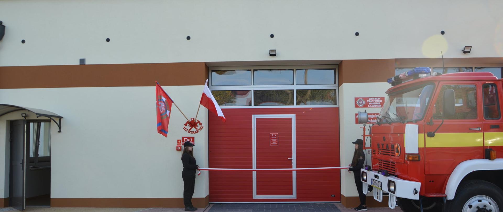 Zdjęcie przedstawia dwie dziewczyny ubrane w mundury koszarowe trzymające biało-czerwoną wstęgo wzdłuż bramy garażowej w zmodernizowanej strażnicy. Przed remizą ustawiony jest samochód ratowniczo-gaśniczy marki Star