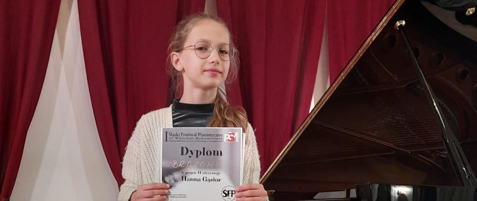 Uczennica stojąca przy fortepianie trzyma w rękach "brązowy" dyplom otrzymany w I Śląskim Festiwalu Pianistycznym im. Władysławy Markiewiczówny w Tarnowskich Górach