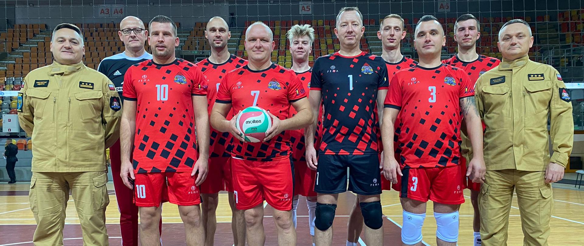 Wspólne zdjęcie grupowe reprezentacji województwa opolskiego strażaków w piłce siatkowej wraz z Opolskim Komendantem Wojewódzkim PSP w Opolu st. bryg. mgr inż. Krzysztofem Kędrykiem oraz Zastępcą Opolskiego Komendanta Wojewódzkiego PSP w Opolu st. bryg. mgr inż. Wojciechem Nawarą. Zdjęcie na boisku do piłki siatkowej znajdujące się w hali. Komendanci w mundurze służbowym koloru piaskowego a reprezentanci w strojach sportowych.