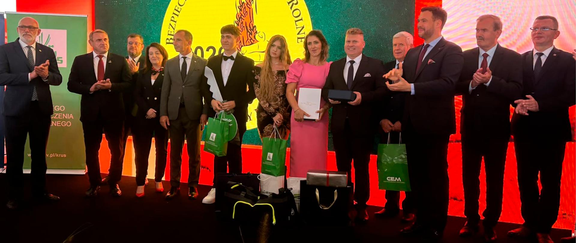 Kilkanaście osób stoi na scenie, w środku stoi rodzina zwycięzców (starszy mężczyzna, kobieta i mężczyzna , nastolatka i nastolatek) Wszyscy trzymają w rękach nagrody, chłopiec trzyma makietę klucza do głównej nagrody – traktora)