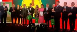 Kilkanaście osób stoi na scenie, w środku stoi rodzina zwycięzców (starszy mężczyzna, kobieta i mężczyzna , nastolatka i nastolatek) Wszyscy trzymają w rękach nagrody, chłopiec trzyma makietę klucza do głównej nagrody – traktora)