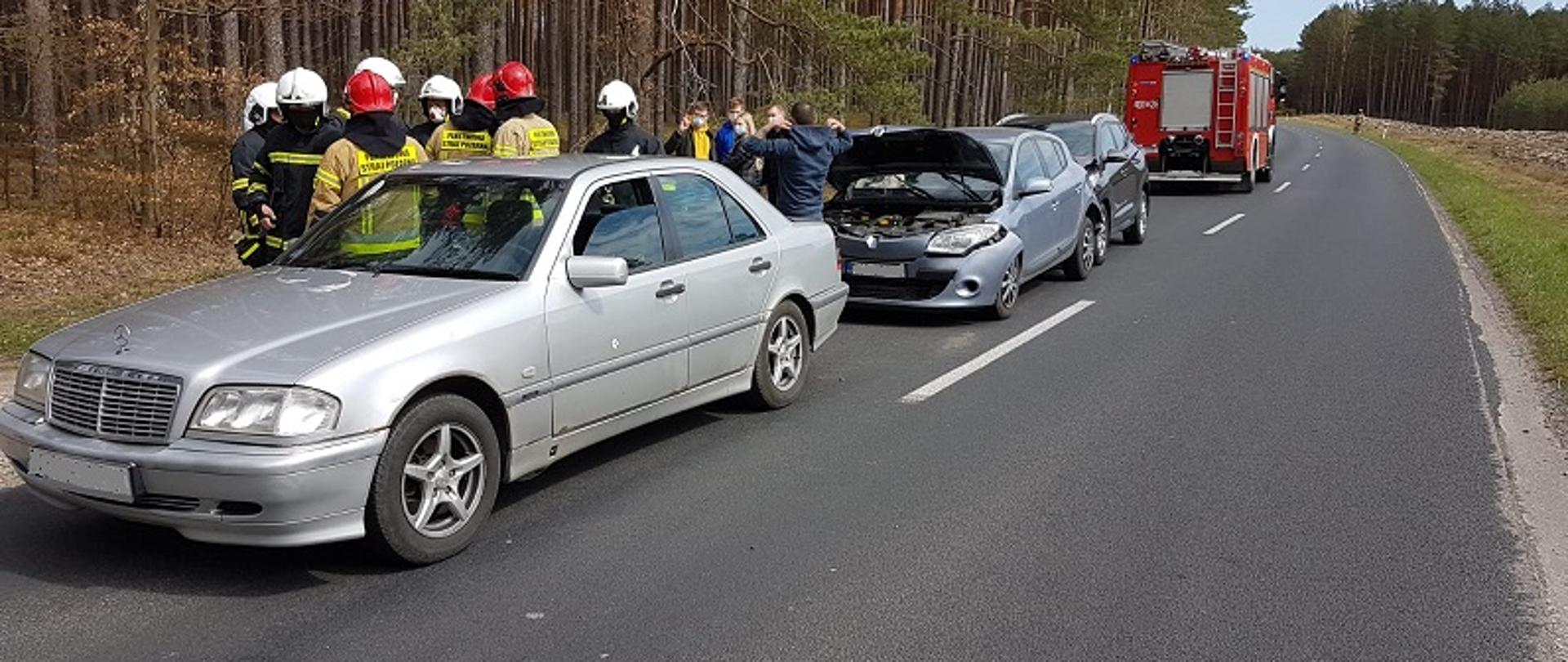 Zdjęcie przedstawia trzy samochody uczestniczące w kolizji na jednym pasie ruchu. Kolejno widoczne od przodu pojazdy Mercedes, Renault i Hyundai wszystkie koloru szarego. Za nimi od czerwony samochód strażacki widoczny od tyłu. Za pojazdami od strony pobocza stoi grupa strażaków i uczestników kolizji
