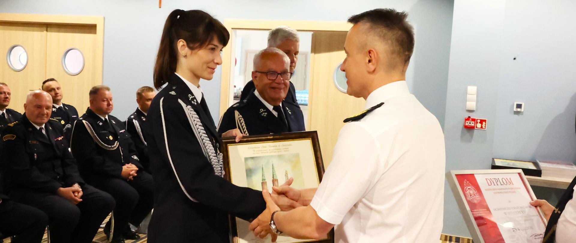 Komendant główny PSP wręcza obraz druhnie OSP, za nią stoi dwóch strażaków