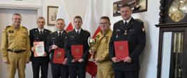 Funkcjonariusze Państwowej Straży Pożarnej stoją obok siebie trzymając czerwone teczki oraz puchar niedaleko ustawiony jest zegar. 