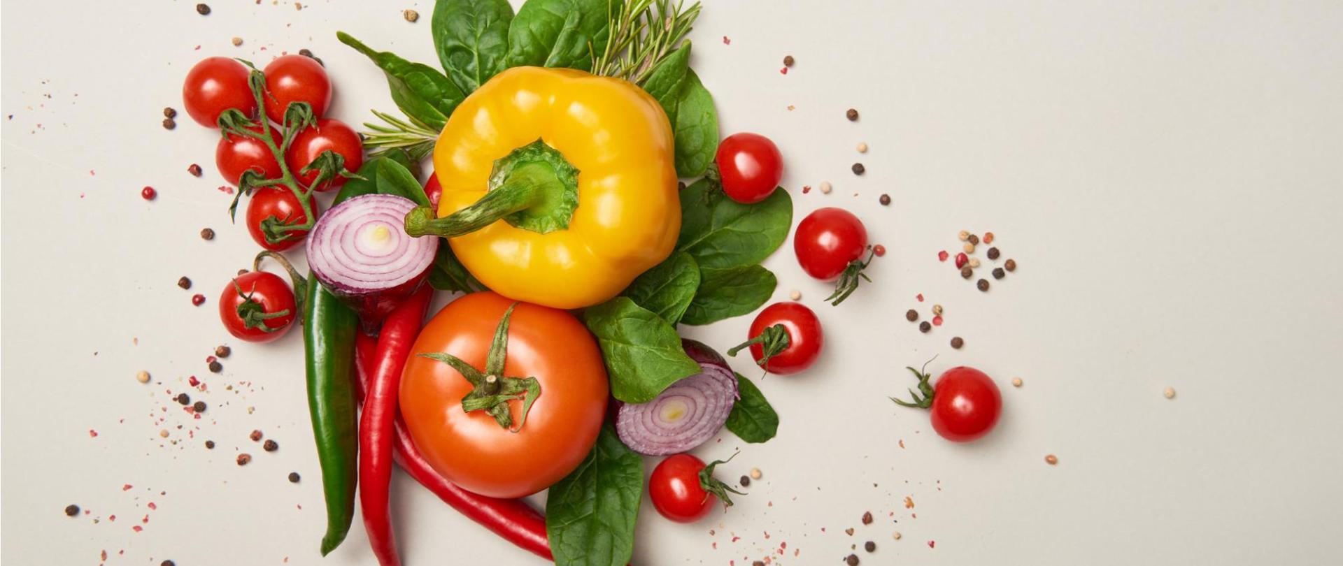 Na zdjęciu na beżowym tle widać warzywa. Wokół nich są ziarenka przypraw.