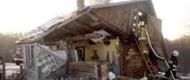 Zdjęcie przedstawia drewniany dom z dobudówką murowaną. Po prawej stronie zdjęcia znajduje się naruszona, drewniana część domu, który uległ uszkodzeniu na skutek wybuchu gazu. Dach domu jest przekrzywiony z powodu uszkodzenia jednaj ze ścian budynku. Po prawej stronie widać strażaka na drabinie, który podaję prąd wody na poddasze w celu ugaszenie tlących się części poddasza. U dołu drabiny widzimy strażaka, odpowiedzialnego za asekurację osoby pracującej na wysokości. Po lewej stronie w oddali widać wóz strażacki. 
