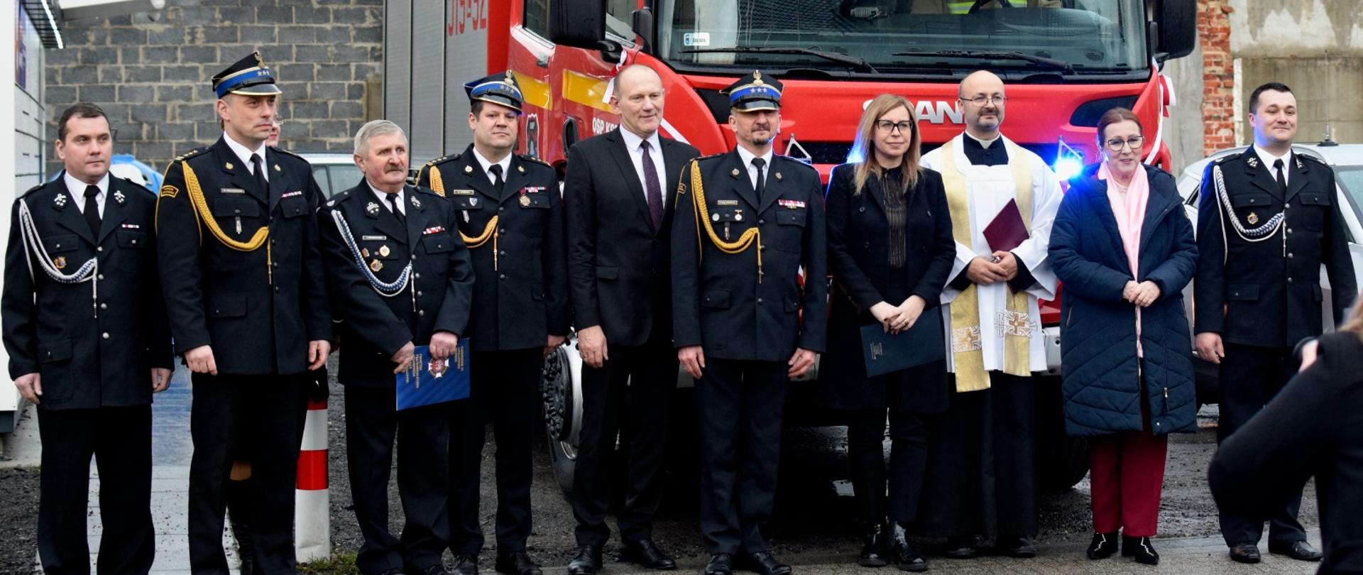 strażacy OSP i PSP oraz dwie kobiety i ksiądz pozują do zdjęcia na tle samochodu gaśniczego