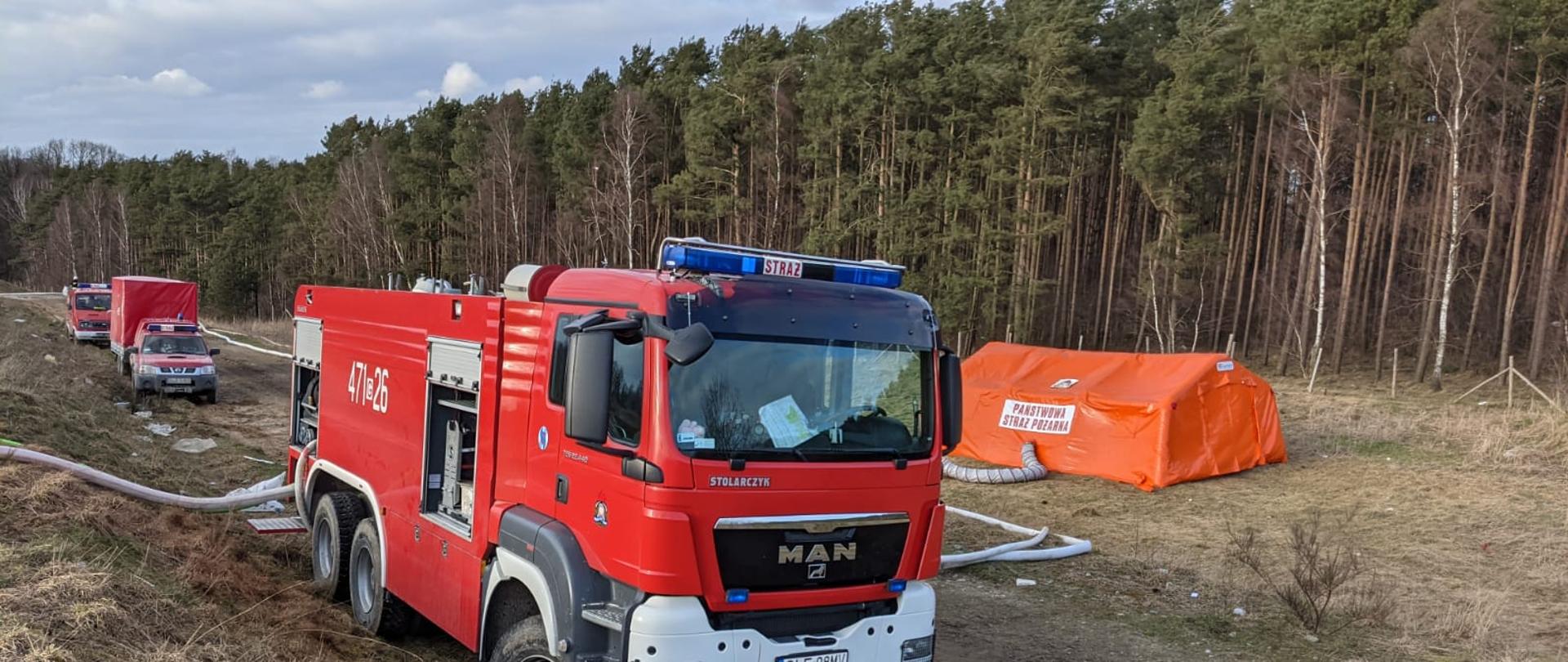 Na zdjęciu na pierwszym planie widać ciężki samochód gaśniczy MAN, który zasilany wodą z lini zasilającej W-110 oraz tłoczy wodę dalej za pomocą lini tłocznej W-110. Za ciężkim samochodem gaśniczym Man stoi lekki saomochód operacyjny Nissan Pickup wraz z przyczepą oraz lekki samochód gaśniczy z motopompą Lublin. Samochody ustawione są na drodze gruntowej. Po prawej stronie na tle lasu poza drogą gruntową rozstawiony jest namiot pneumatyczny. 