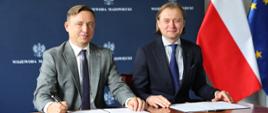 Prezes PAA Andrzej Głowacki i Wojewoda Mazowiecki Mariusz Frankowski podczas podpisania listu intencyjnego w sprawie monitoringu radiacyjnego. Prezes PAA trzyma w ręku długopis. W tle flagi Polski i Unii Europejskiej