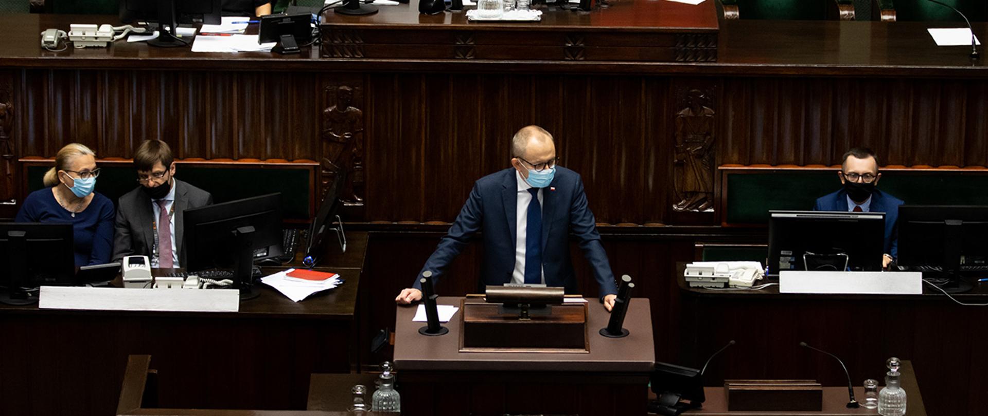 Wiceminister Błażej Poboży przemawiający z mównicy sejmowej. Po prawej i po lewej stronie protokolanci sejmowi.