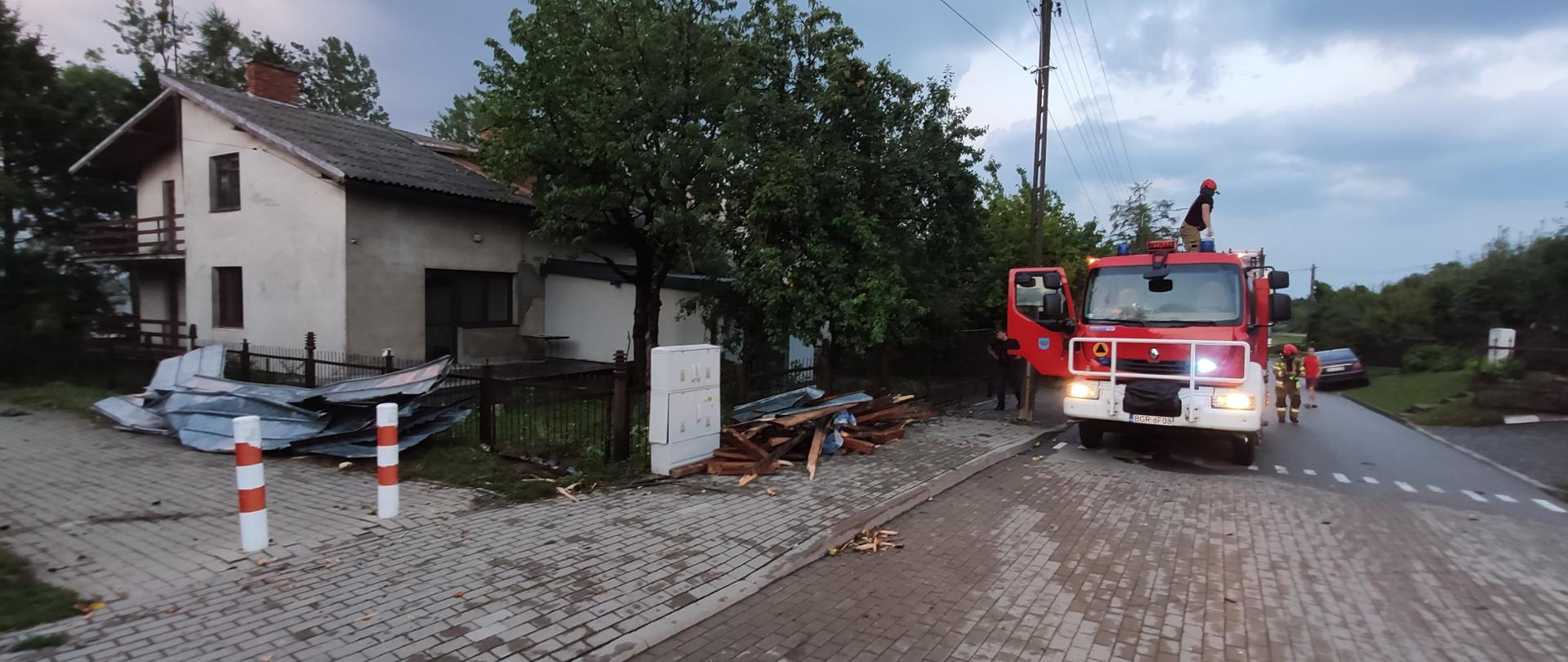 Samochód strażacki stoi na ulicy. Przy budynku leży częściowo zerwany dach.