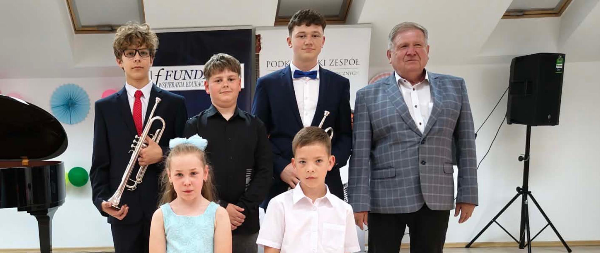 Grupa uczniów oraz nauczyciel szkoły muzycznej pozują do pamiątkowego zdjęcia na scenie sali koncertowej. Wśród uczniów znajdują się młodzi instrumentaliści, w tym dwaj trębacze, a po lewej stronie widoczny jest fortepian koncertowy. Uczestnicy występu ubrani są w stroje galowe, a zdjęcie wykonano podczas szkolnego wydarzenia artystycznego.