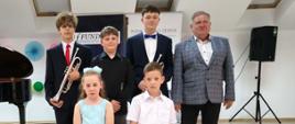 Grupa uczniów oraz nauczyciel szkoły muzycznej pozują do pamiątkowego zdjęcia na scenie sali koncertowej. Wśród uczniów znajdują się młodzi instrumentaliści, w tym dwaj trębacze. Uczestnicy występu ubrani są w stroje galowe, a zdjęcie wykonano podczas szkolnego wydarzenia artystycznego.