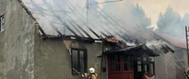 Na zdjęciu widać budynek mieszkalny ze spalonym dachem oraz strażaków, którzy podają wodę do jego wnętrza.