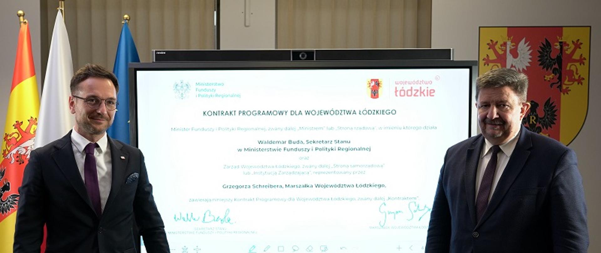 Wiceminister Waldemar Buda i marszałek Grzegorz Schreiber stoją na tle interaktywnej tablicy, na której widać podpisany kontrakt programowy dla woj. łódzkiego