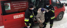 Na zdjęciu widać dwóch ratowników OSP. Jeden ratownik trzyma kobietę w podeszłym wieku za łokieć a drugi stoi po lewo kadru i jej asystuje stojąc obok. Kobieta wyszła z auta i stoi na ziemi. Jest ubrana w zimową kurtkę jasnej barwy, czarną spódnicę, spodnie barwy czarnej oraz czapkę koloru fioletowego, na twarzy ma maseczkę jednorazową. W prawej ręce trzyma małą torebkę. Ratownicy są ubrani w ubranie typu Nomex z odblaskami barwy jasnożółtej i szarej, na głowie mają czapkę wełnianą barwy czarnej, maseczkę na twarzy oraz rękawiczki jednorazowe. W tle samochody osobowe. Na ziemi widać śnieg. Auto jest typu furgon barwy czerwonej z napisem na prawych przednich drzwiach OSP Stary Waliszew, po lewej stronie auta widnieje numer operacyjny samochodu oraz napis Straż.