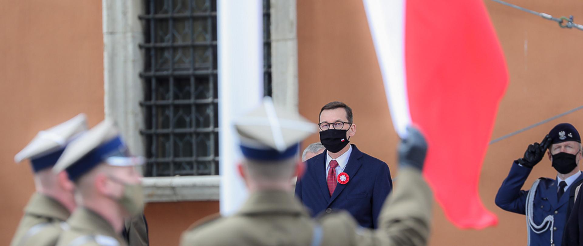 Premier Mateusz Morawiecki wpatruje się we flagę wciąganą na maszt.