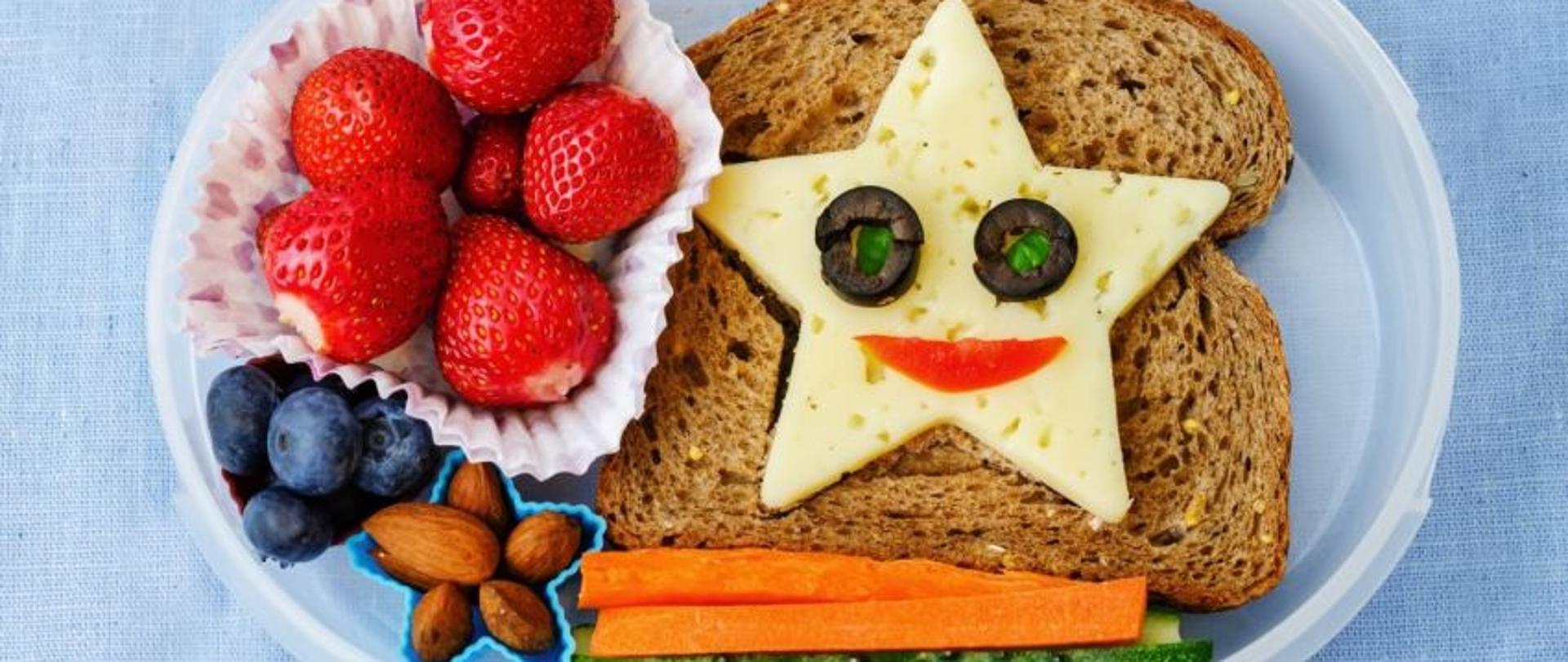 na zdjęciu pojemnik z jedzeniem. kanapka z serem wyciętym w gwiazdę, w środku sera oczy z oliwek, obok truskawki, borówki, migdały, marchewka i ogórek