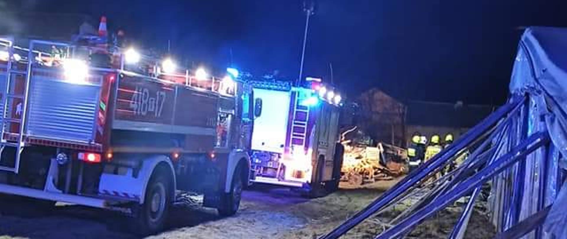 Zdjęcie przedstawia po lewej stronie dwa samochody ratowniczo-gaśnicze gaśnicze z włączoną alarmową sygnalizację świetlną . Na drugim planie znajduje się kilku strażaków w ubraniach specjalnych. Po prawej stronie widnieje część tunelu foliowego podpartego drewnianymi belkami. Zdjęcie zrobione jest w porze nocnej.