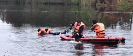 Na rozmarzającej tafli jeziora utrzymuje się w wodzie jedna osoba, w jej kierunku na desce lodowej płynie trzech ratowników w kamizelkaza z napisem STRAŻ na plecach.