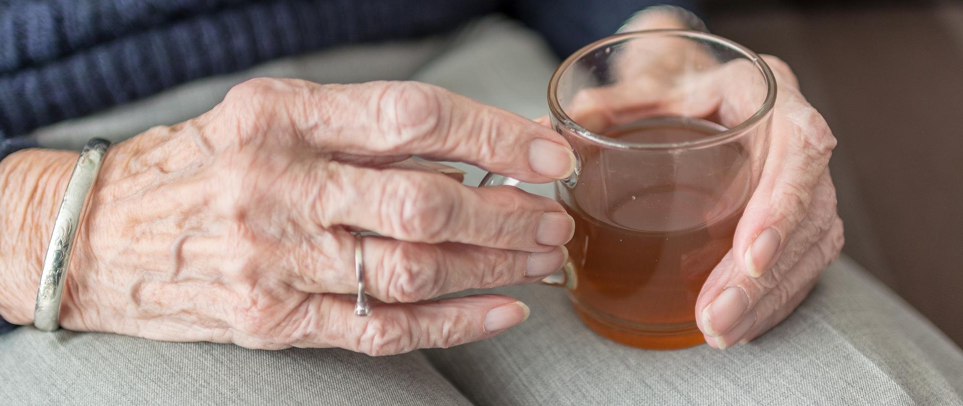 Na fotografii dłonie starszej kobiety trzymającej kubek z gorącą herbatą. 