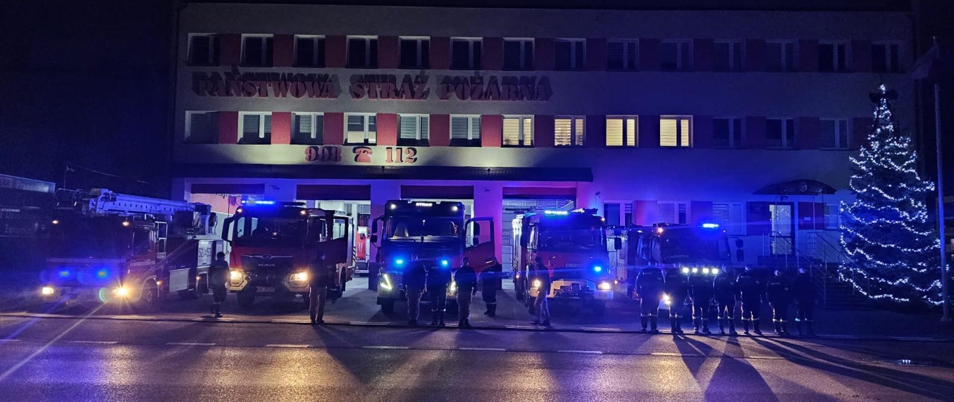 Na zdjęciu widać pojazdy straży pożarnej zaparkowane przed budynkiem Komendy Powiatowej Państwowej Straży Pożarnej. Strażacy stoją w szeregu, ubrani w mundury, a wokół nich migają niebieskie światła sygnałowe. W tle widać choinkę ozdobioną światełkami, a na budynku widnieje napis „PAŃSTWOWA STRAŻ POŻARNA”. Scena ma spokojny, ale jednocześnie poważny charakter, oddając hołd strażakowi.
