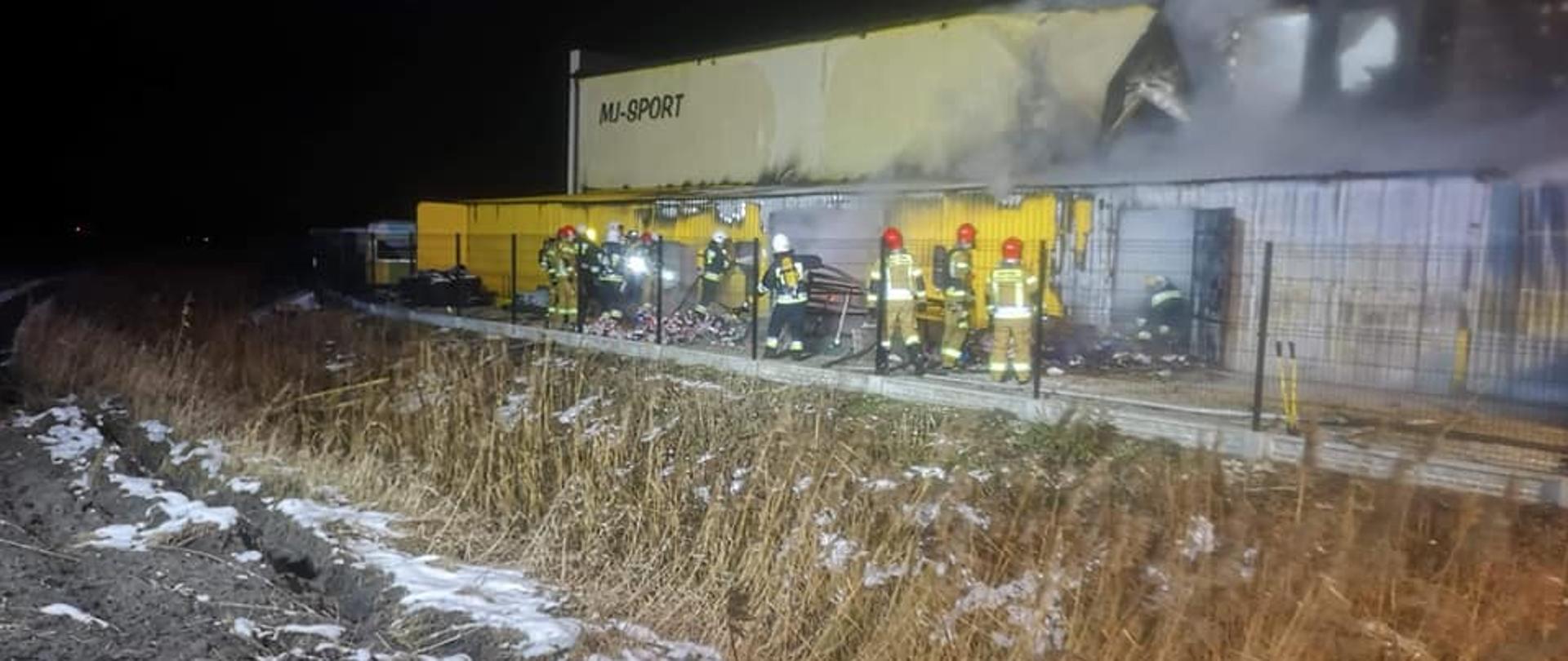 Pora nocna. Na pierwszym planie pole porośnięte wysoką suchą trawą. Lekko ośnieżone. W oddali metalowy żółty budynek przylegający do jasnego wyższego budynku. Dużo dymu wydobywa się z jego górnej części. Kilkunastu strażaków gasi pożar.