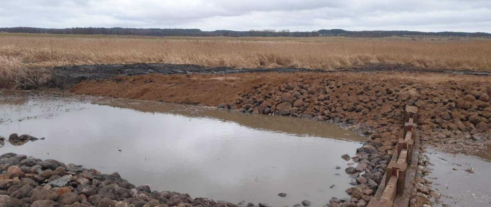 Na zdjęciu znajduje się obiekt małej retencji wodnej - próg piętrzący ze szczelnymi ściankami wykonany na rzece Wąż na terenie Bagna Nietlickiego, stanowiącego jedną z enklaw obszaru Natura 2000 Mazurskie Bagna. W dalszej części zdjęcia widoczne są podmokłe siedliska przyrodnicze, chronione w ramach tego obszaru Natura 2000.