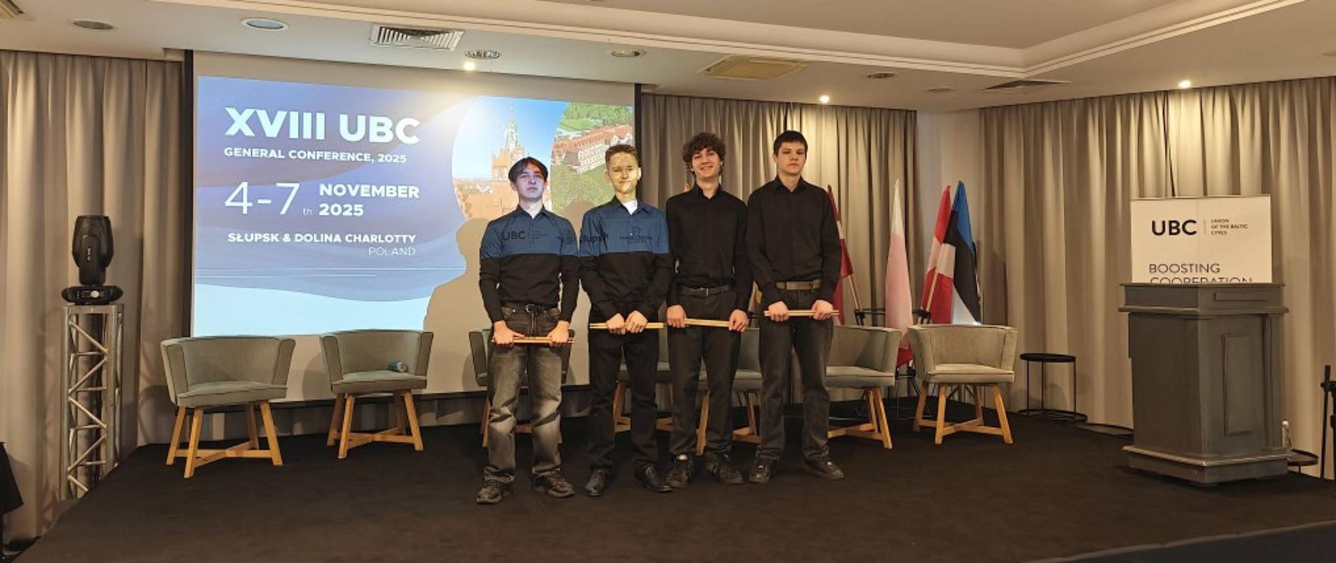 Zdjęcie kolorowe, czterech chłopców stojących na czarnej scenie wewnątrz sali konferencyjnej, z tyłu duży ekran z napisem "XVIII UBC General Conference 2025 4-7 November in 2025 Słupsk & Dolina Charlotty Poland", z lewej i prawej strony ekranu dwa jasnoszare fotele, po prawej stronie czarna mównica z logo UBC, zasłony w oknach w tle.