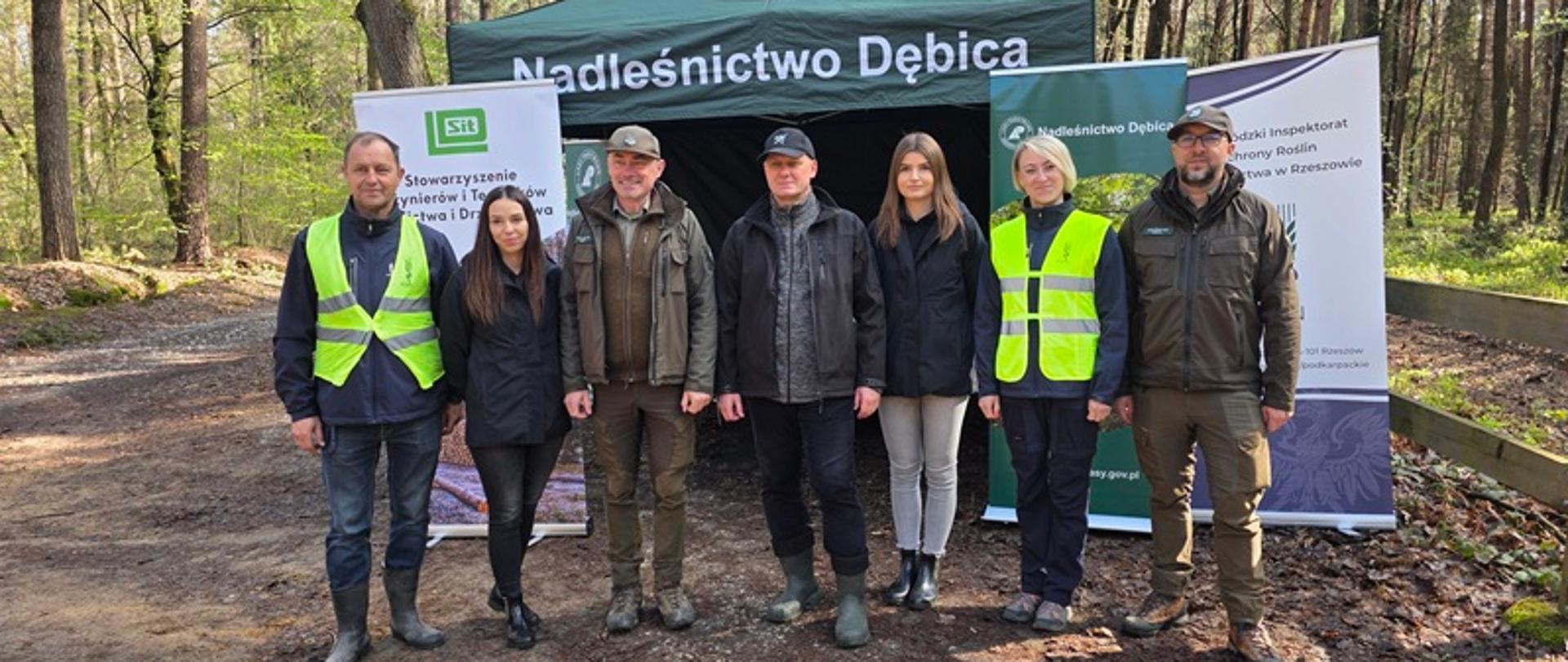 Siedem osób stoi w rzędzie na leśnej ścieżce przed zielonym namiotem z napisem „Nadleśnictwo Dębica”. Część z nich ma na sobie odblaskowe kamizelki w kolorze neonowej zieleni, inni ubrani są w kurtki terenowe i gumowe buty. Po obu stronach namiotu ustawione są roll-upy z dodatkowymi informacjami i logotypami instytucji związanych z leśnictwem. Tło stanowi las z wysokimi drzewami i miękkim, naturalnym światłem dziennym. Grupa pozuje do zdjęcia, patrząc w stronę aparatu.