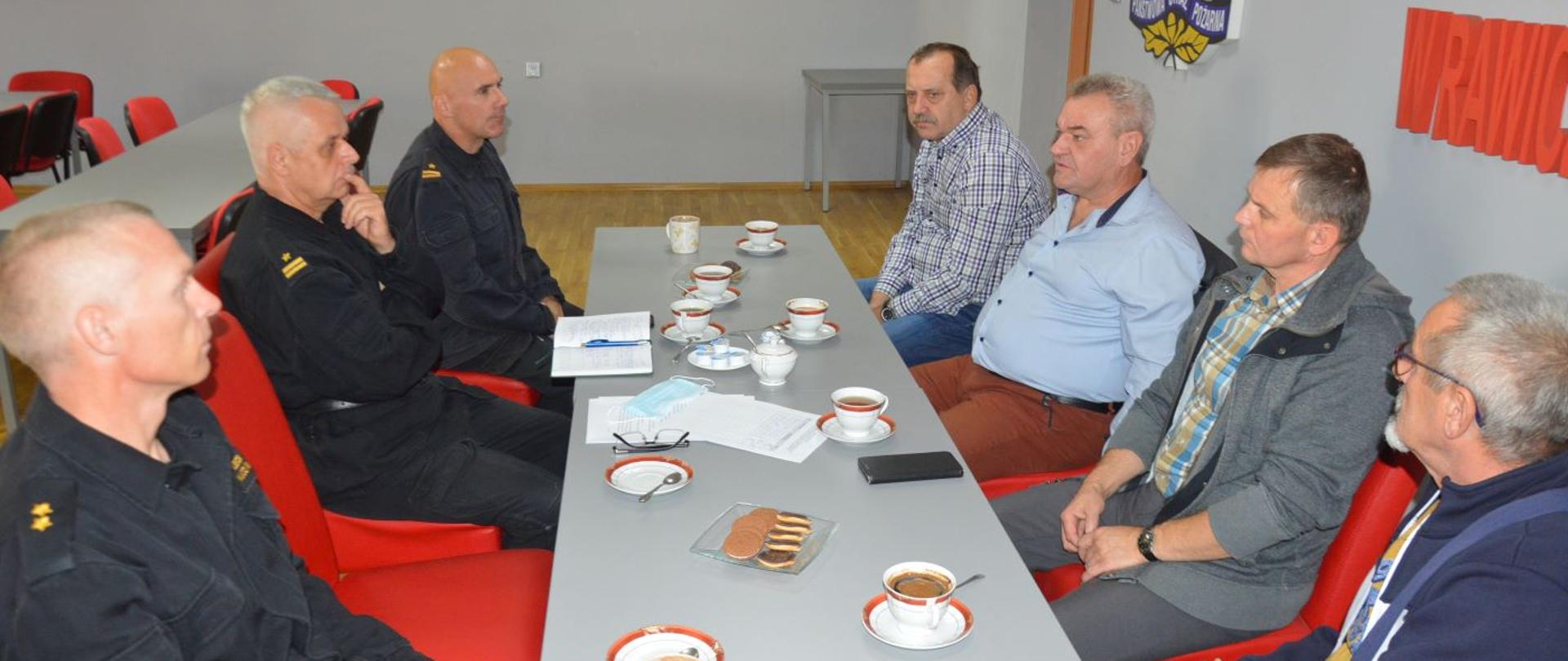 Narada z kominiarzami i zarządcami budynków wielorodzinnych. Świetlica KP PSP w Rawiczu. Przy wspólnym stole siedzi trzech funkcjonariuszy komendy, a na przeciwko nich czterech mężczyzn . Dwaj z nich to mistrzowie kominiarscy, pozostali - zarządcy budynków wielorodzinnych w Rawiczu. W tle na ścianie logo PSP i napis "Komenda Powiatowa PSP w Rawiczu" Na stole filiżanki z kawą, ciastka, talerzyki.